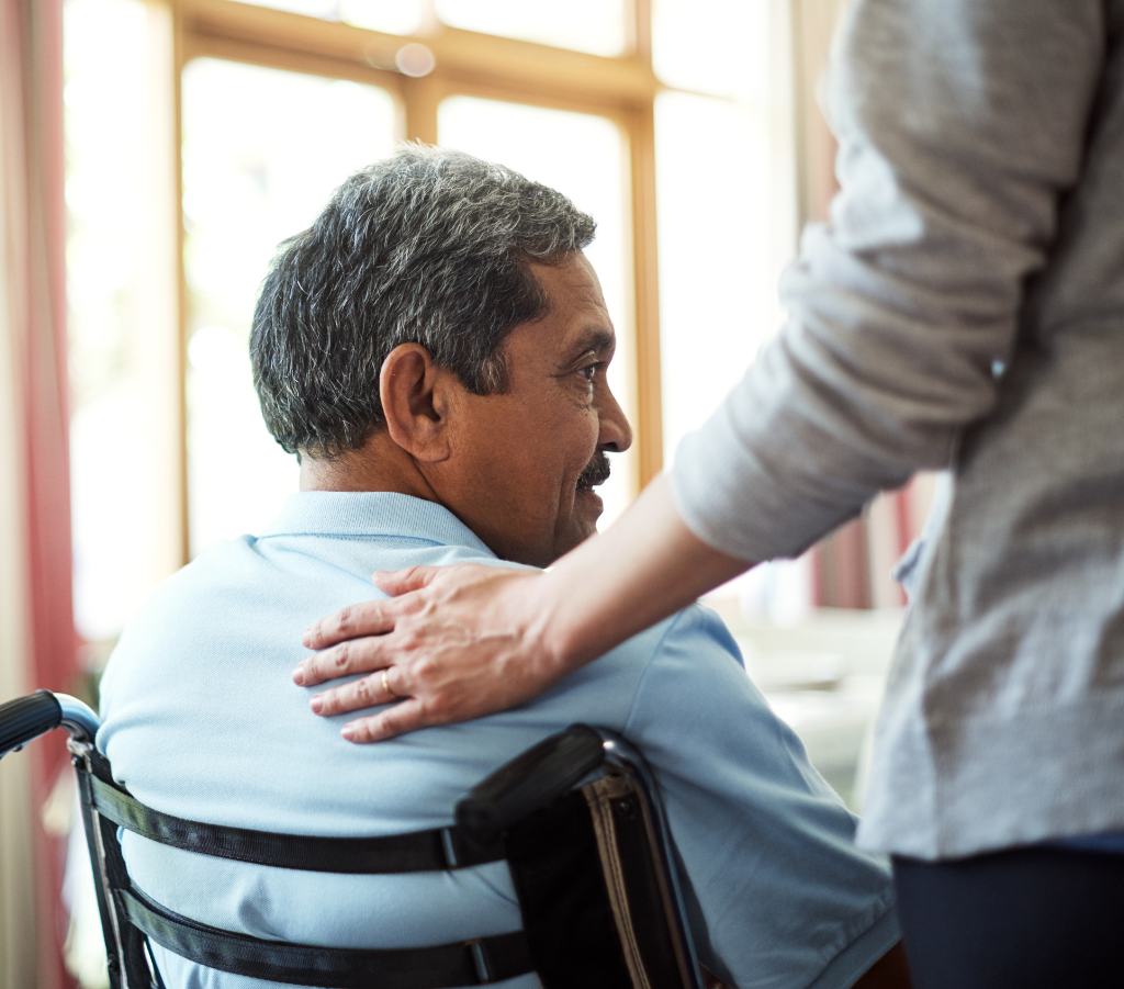 This image depicts a person comforting a man in a wheelchair by placing their hand on his right shoulder