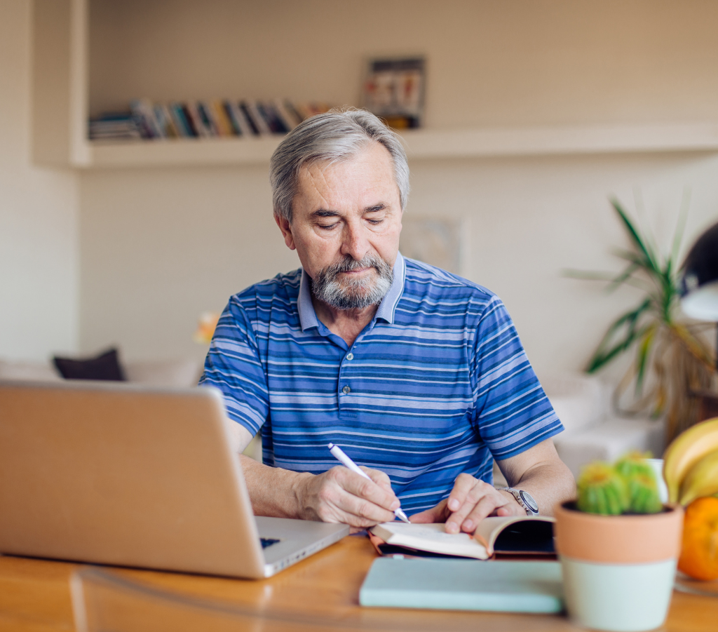 This image depicts an older man writing in a notebook whilst sitting in front of a laptop computer
