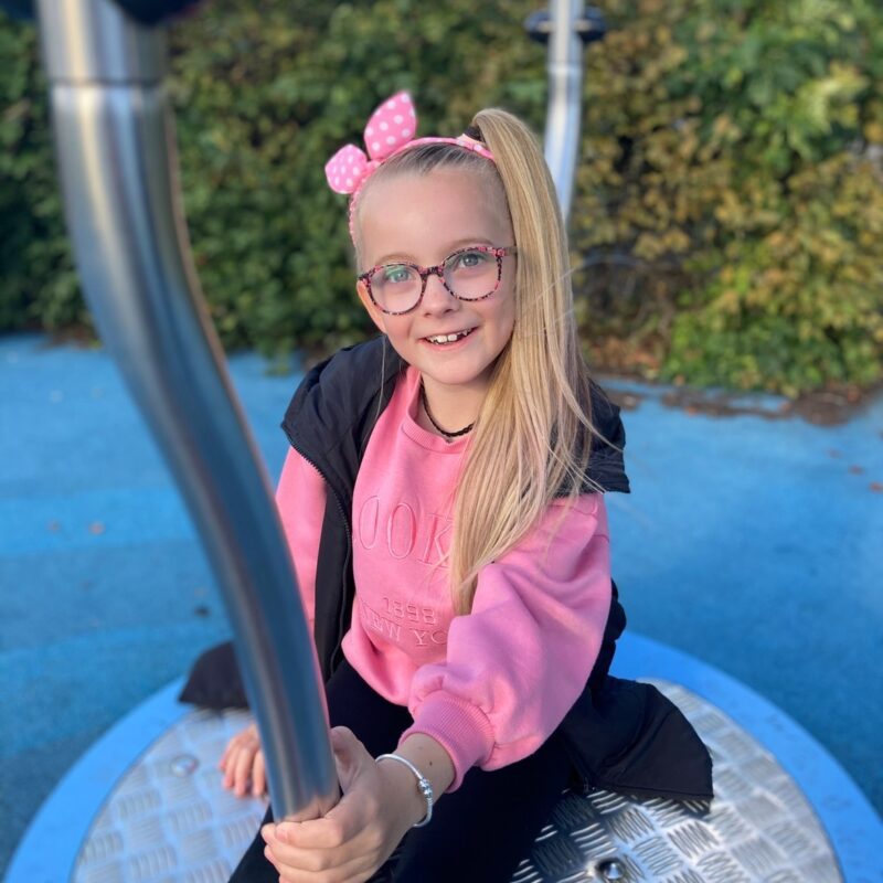 Sienna sits on playground ride, smiling at the camera. She has blonde hair, glasses, and has a pink headband and jumper.