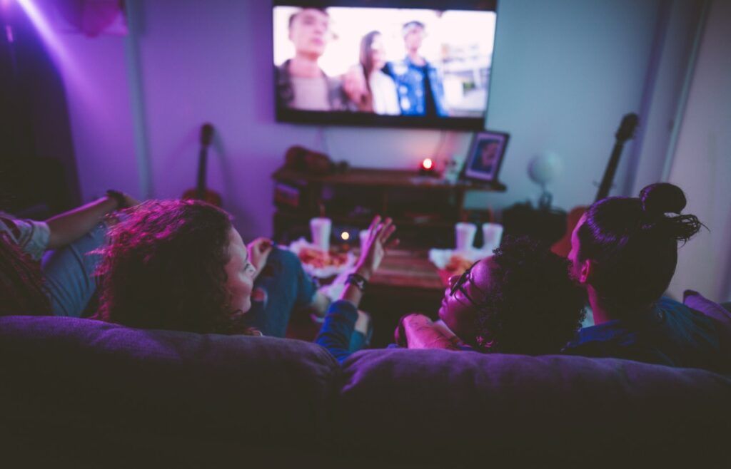 A group of young adults sitting on a couch watching TV with takeaway food on the table in front of them