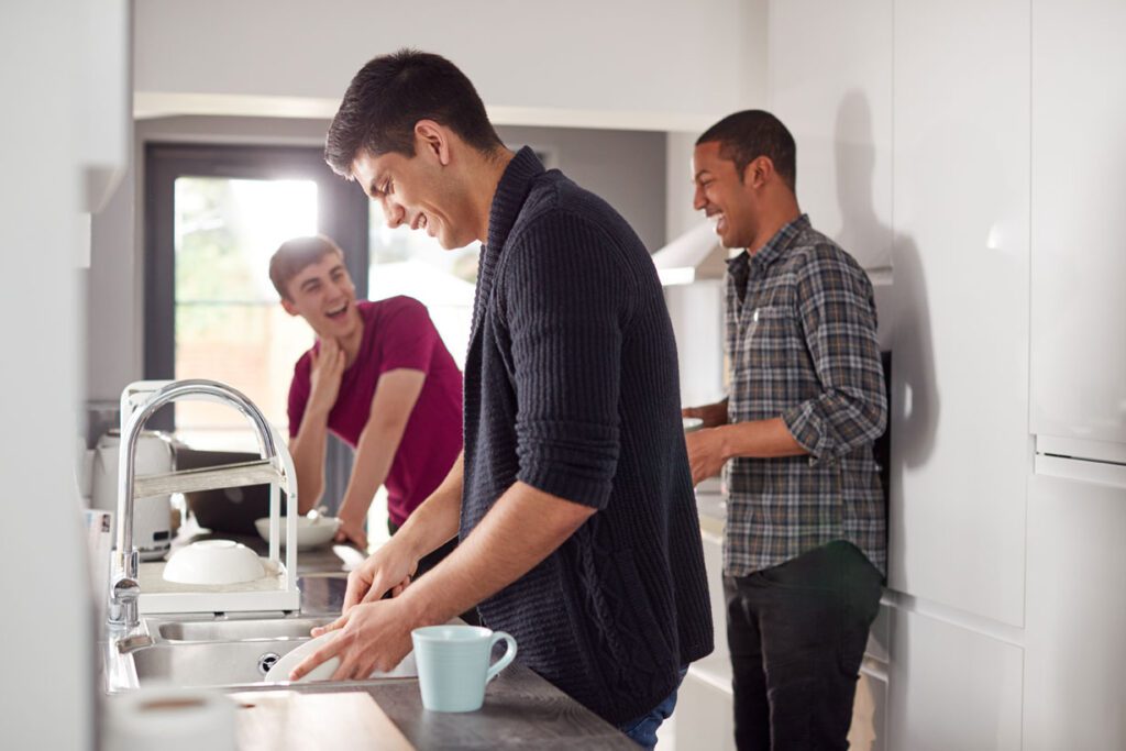 A young man washing a dish in a kitchen sink while two other people talk to each other behind him.