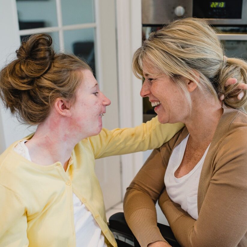 a young woman in a wheelchair holding onto an older woman who is crouched down. Both are laughing.