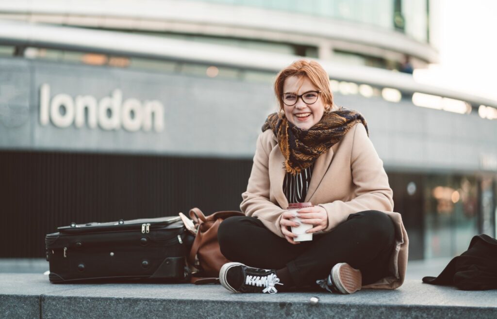 a young woman sitting with a coffee and luggage outside a london station